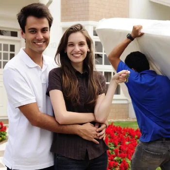 a couple holding the key to a new house and a mover in the background carrying a mattress